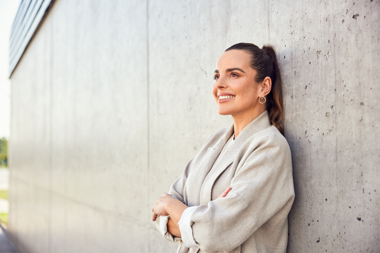 A woman in a blazer leaning up against a wall smiling